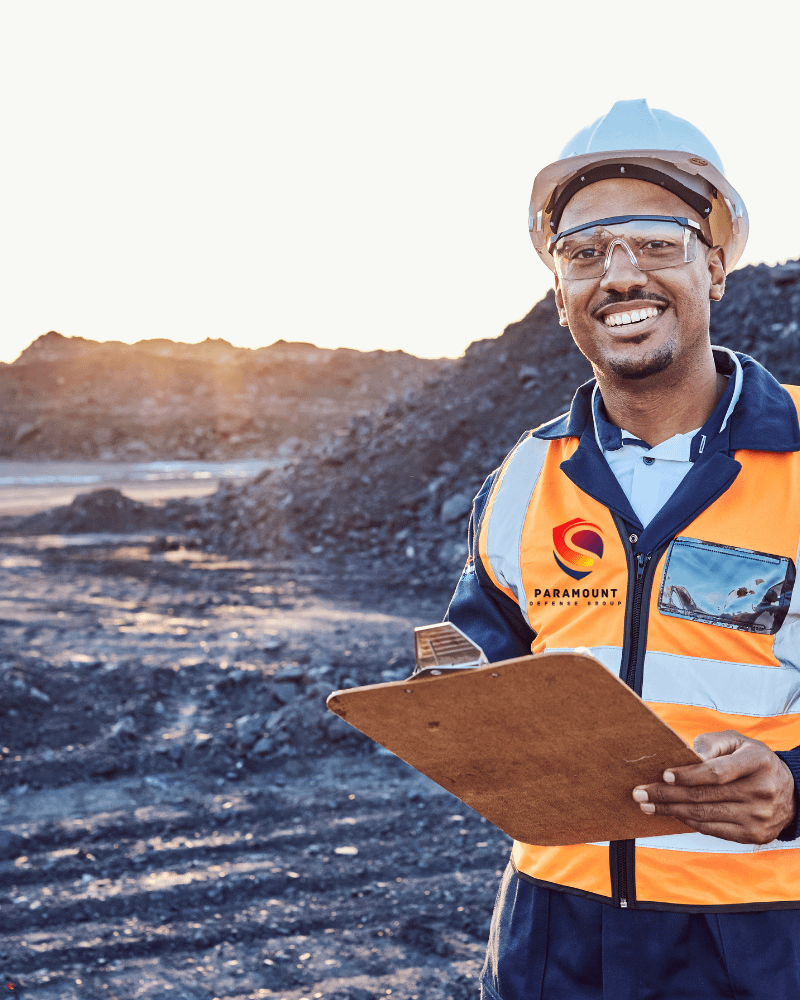 A smiling industrial worker wearing safety gear, including a hard hat, safety glasses, and a high-visibility vest, holding a clipboard at a mining or construction site with piles of excavated material in the background.